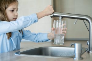 Young girl in blue shirt filling a glass with tap water from the kitchen sink